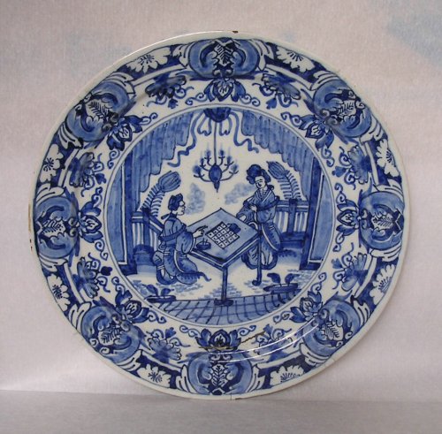 Plate with blue design on white ground. The central part of the plate shows two figures in Chinese-style clothing playing a board game at a table. The figure on the right is standing, while the one on the left is kneeling. Above them hangs a chandelier. There is a repeating floral border round the edge of the plate. 