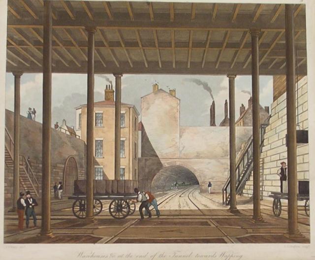 The viewer is inside a warehouse building looking out at railway tracks heading into a tunnel. In the mid-ground, two men push a cart laden with goods towards the left. To the far right, another man stands on an empty cart. Buildings rise above the tunnel with smoking chimneys. 