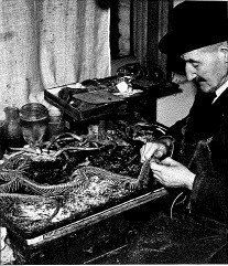 Black and white photograph of a male staff member in a trilby skinning and cleaning a snake skeleton. He sits at a desk which has his tools and other unfinished taxidermy works around him.