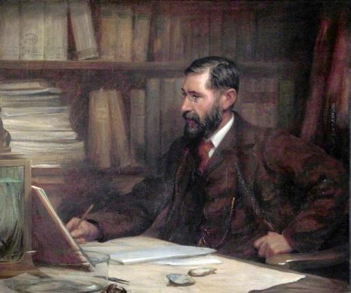 Portrait of a man sat at a desk. He is studying with a book open next to him and some oyster shells to his left. He wears a tweed suit, matching waistcoat, white collar and red tie. He has dark brown hair neatly cut and a beard.He has thin rimmed spectacles on, there is a library of books behind him. 