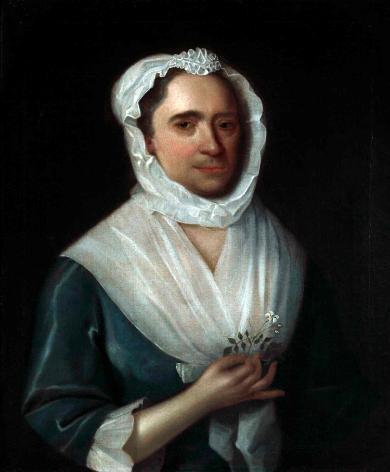 Portrait of a woman wearing a blue silk dress holding a jasmine flower with a white bonnet and collar. She holds a jasmine flower in her right hand. The lower half of her face has a five o'clock shadow. 