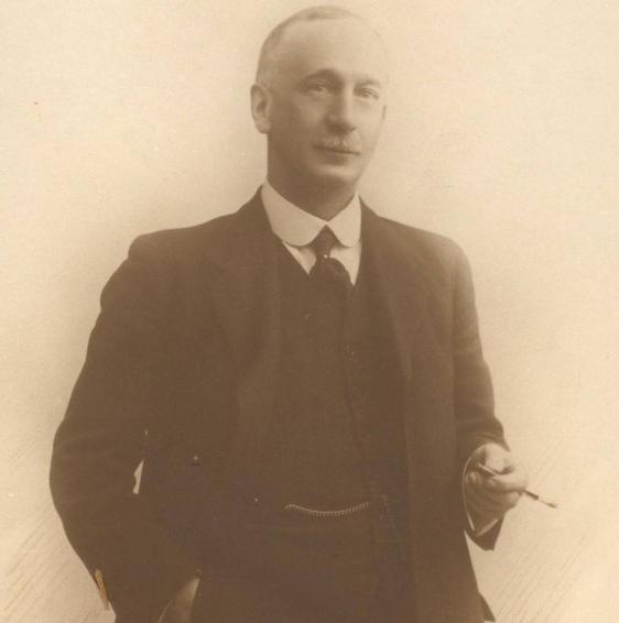 Sepia photograph of the Professor Andrew Melville Paterson.He is dressed in a smart suit, starched round collar and tie. He has a grey moustache and thinning grey hair.He stands with his right had in his pocket, holding a cigarette in a holder in his left hand. He half smiles.