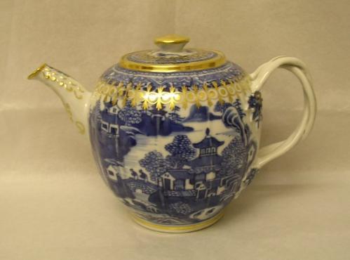 Teapot with lid and twist-style handle. The body has a blue design on white ground with a landscape scene showing pagoda-style buildings and trees set against water. There are two figures on a bridge. The teapot has gilding on the lid, spout and around the top of the body. 