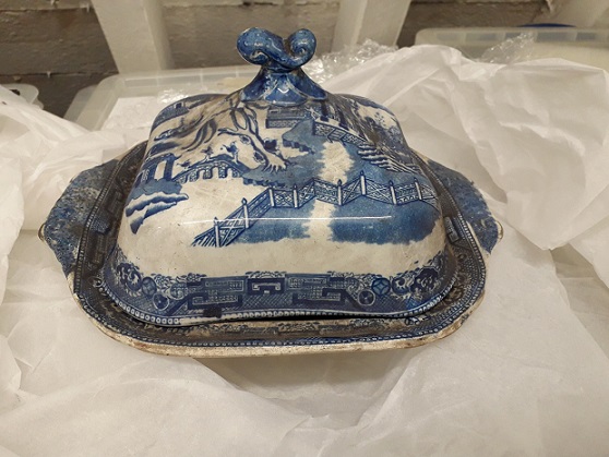 Lipped dish with domed cover in blue and white Willow Pattern design. It is rather dusty. The dish has been photographed on white tissue paper with storage cases in the background. 