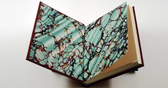 A hard-backed book is held open. Inside is blue and brown marbled paper.