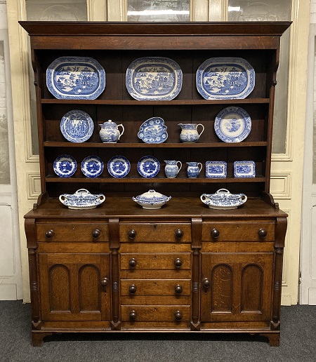 A wooden kitchen dresser with three shelfs at the top and drawers and cupboards underneath. Blue and white tablewares are set out on the shelves, with three large Willow Pattern plates on the top shelf. The plates are set upright so they face outwards with the pattern showing.